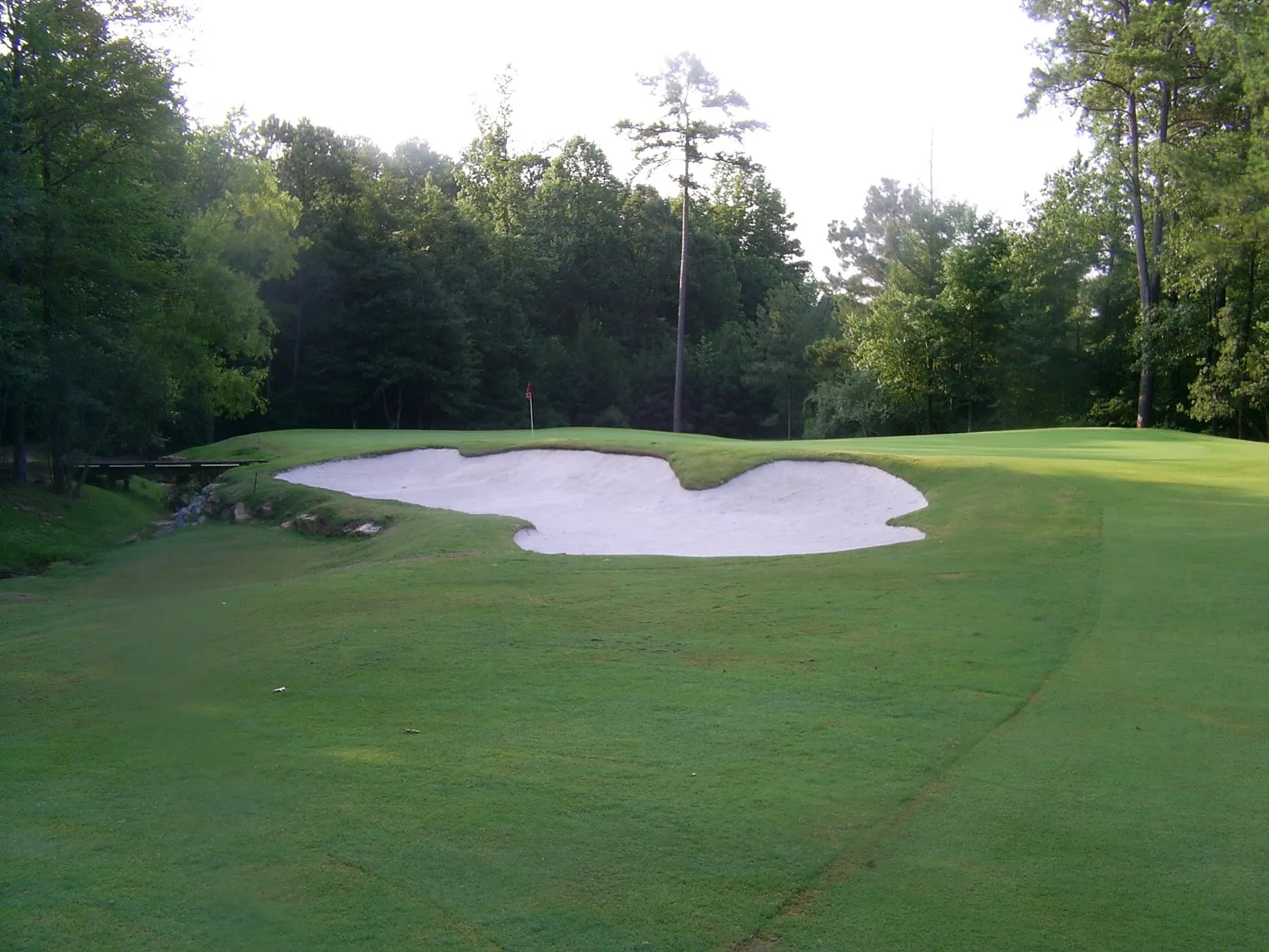 A white sand bunker set into a mown green surround, with a small bridge over a creek at the left and hardwoods behind.