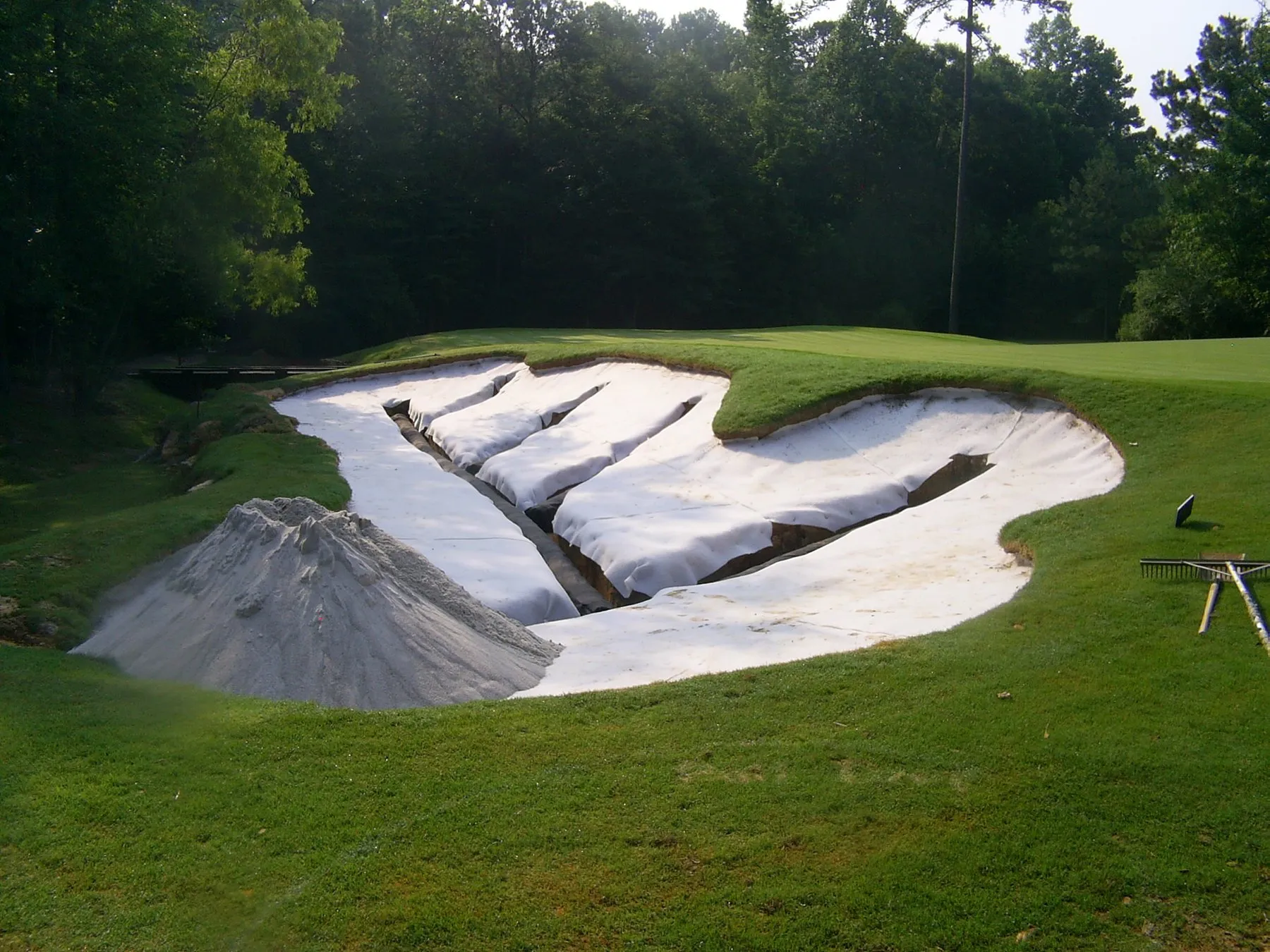 A bunker lined with white fabric over its drainage, sand piled ready to spread, with a finished green and a creek behind.