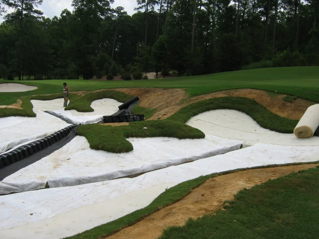 Black drainage pipe and white liner laid out across two newly shaped bunkers, a roll of sod at the edge and a worker in the distance.