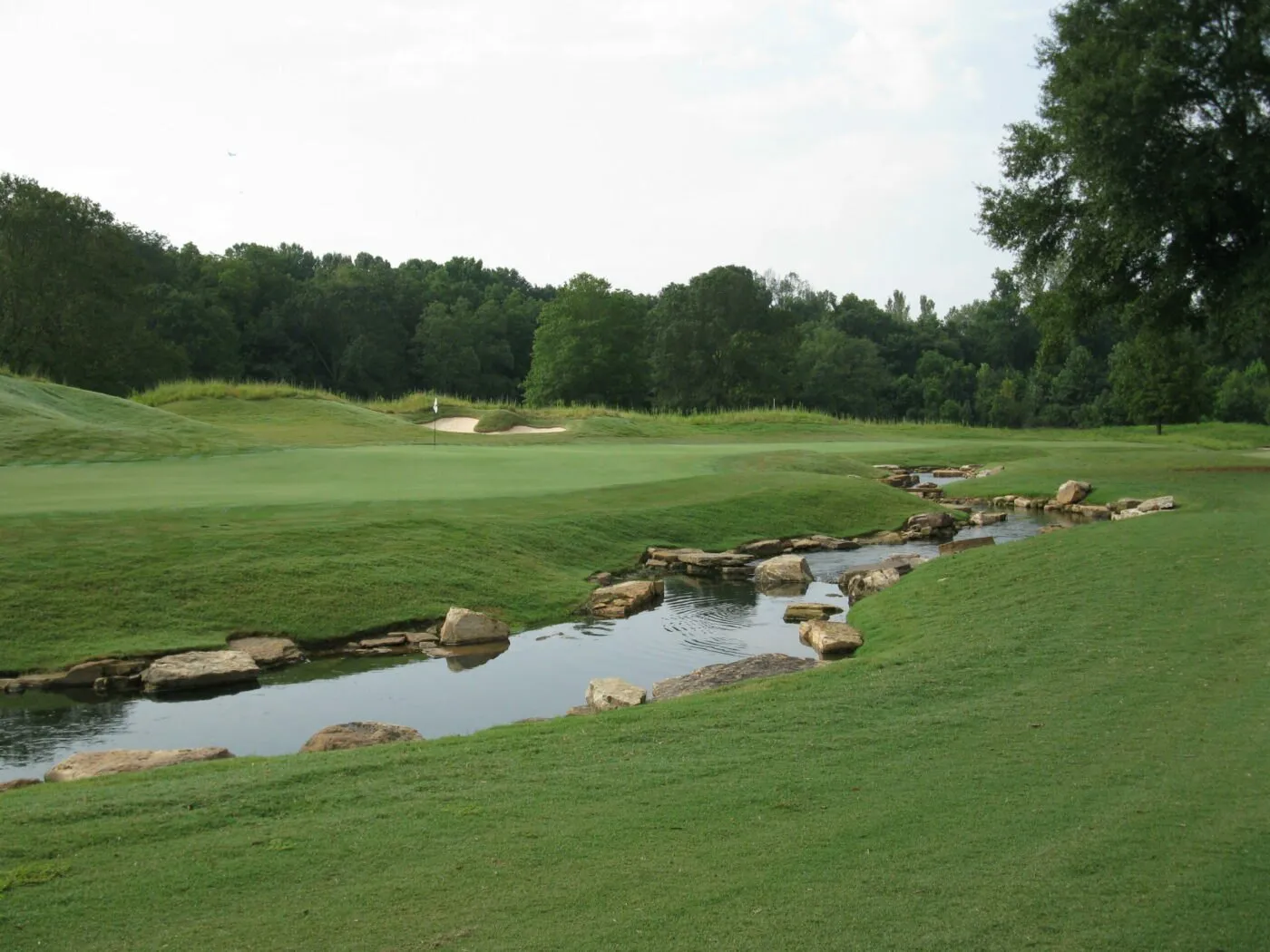A rock-lined stream running past a green and its bunker, with a wooded ridge behind.