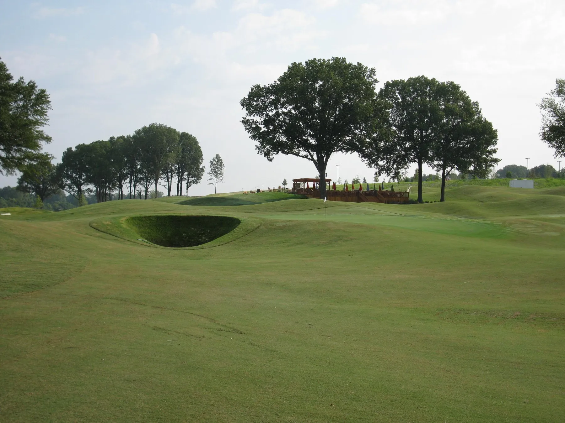 A deep grass-walled bunker cut into a mown slope, with a spreading oak and the clubhouse terrace on the ridge behind.