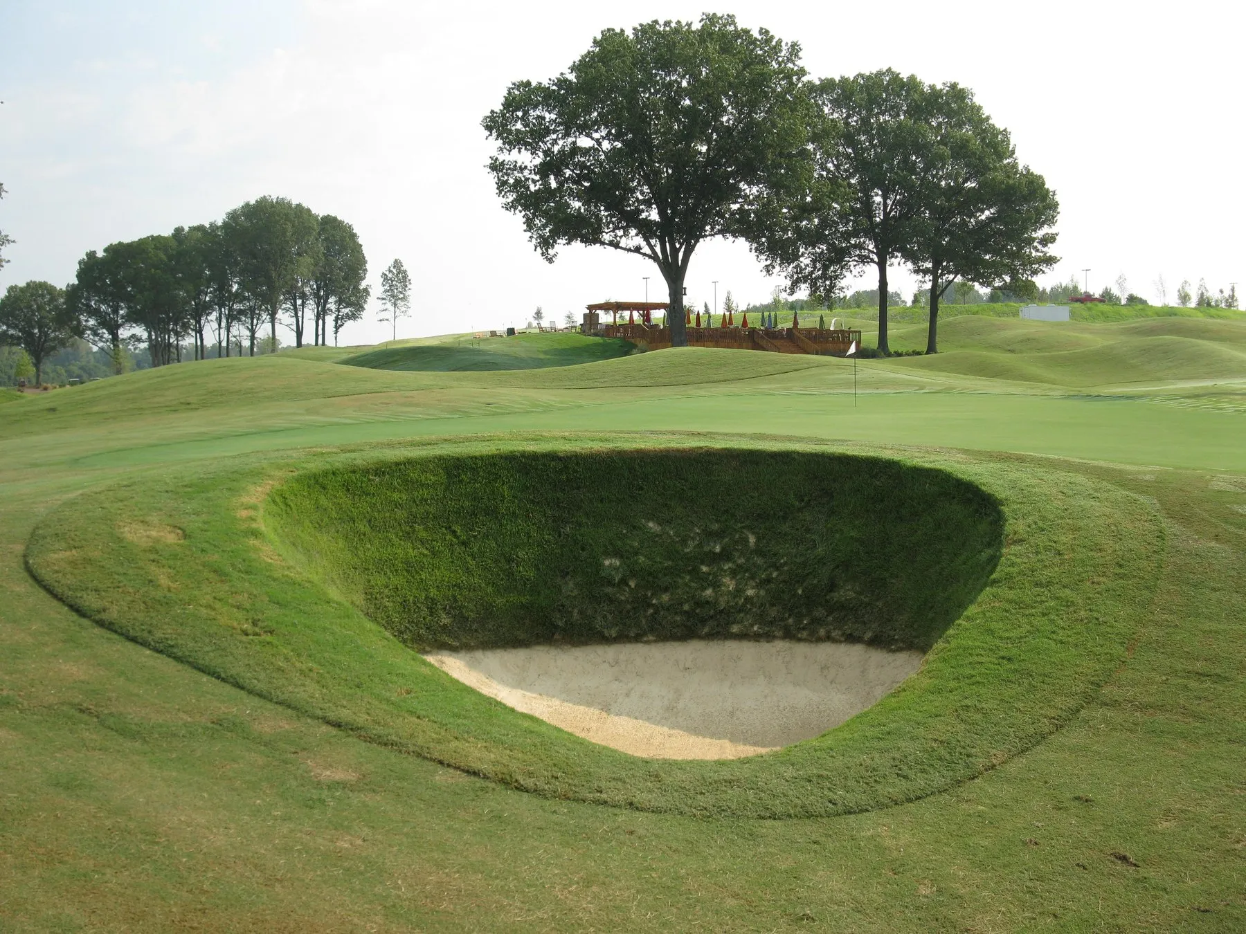 A deep grass-walled bunker with a narrow sand floor, cut into mown turf below a spreading oak.