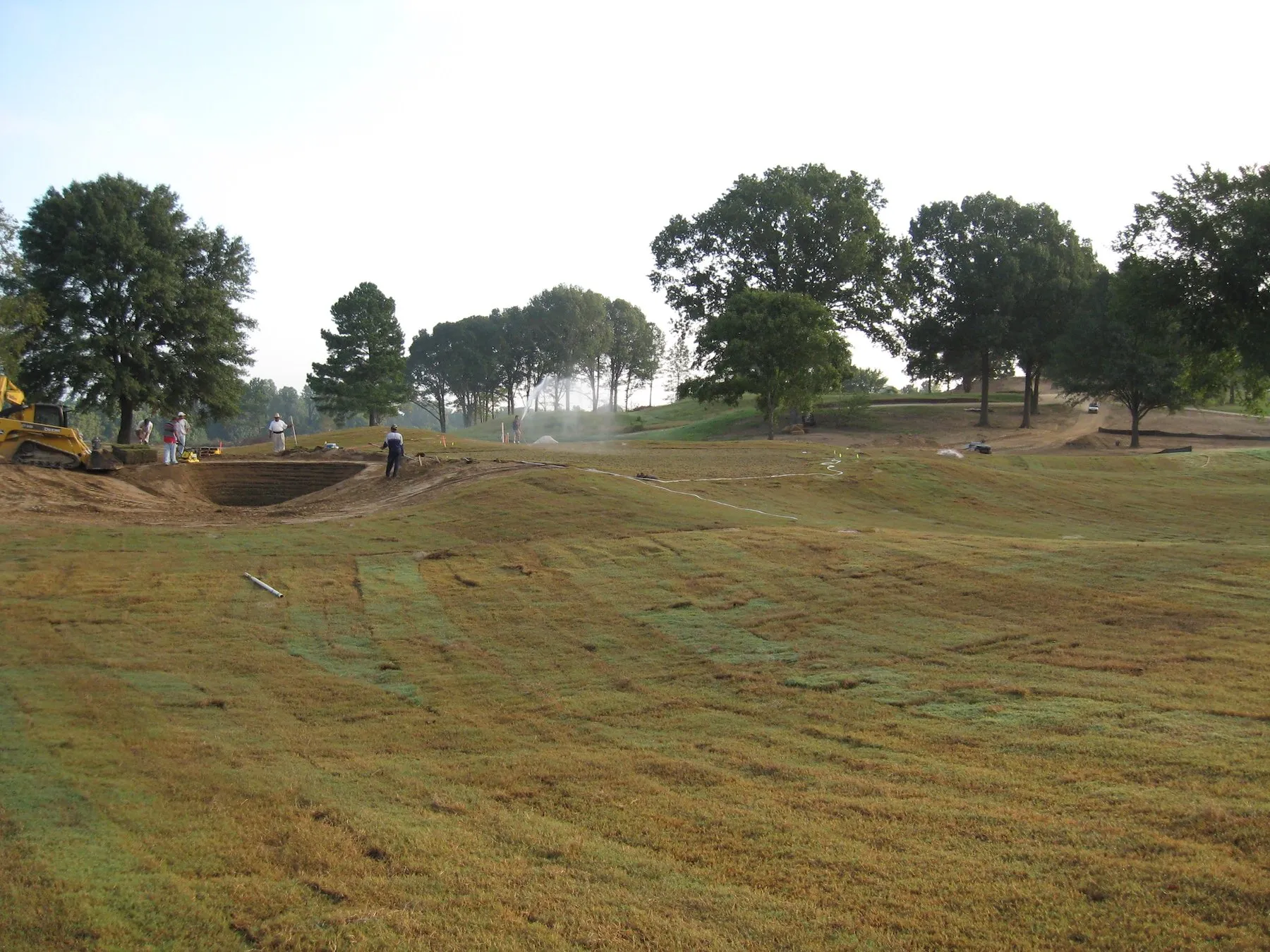 A newly shaped and sprigged fairway sweeping up toward a line of mature trees, with a crew and a track loader working at a fresh bunker on the left.