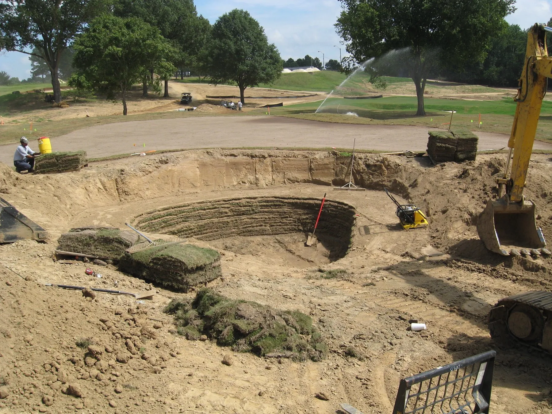 The stepped base of a bunker under construction at Mirimichi, rolls of sod stacked at the edge and an excavator alongside.
