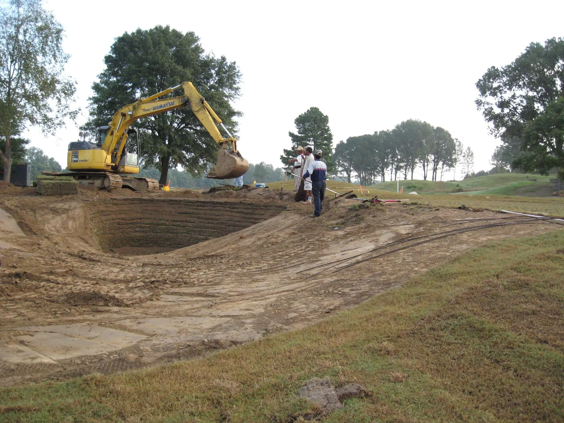 An excavator shaping a large bunker at Mirimichi while two men look on from the rim.