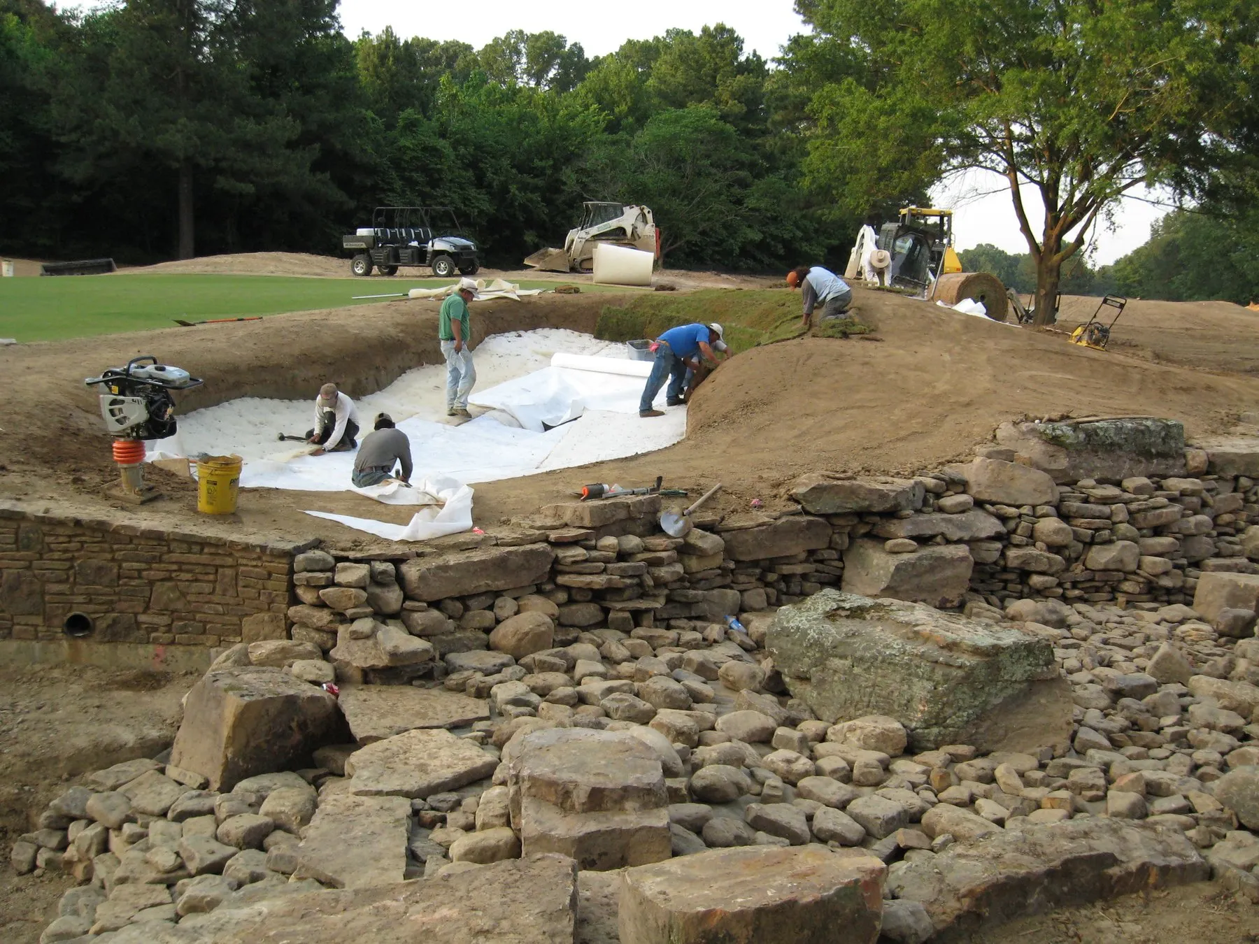 A crew laying liner in a bunker under construction above a dry-stone wall, with a skid steer and carts on the grass behind.