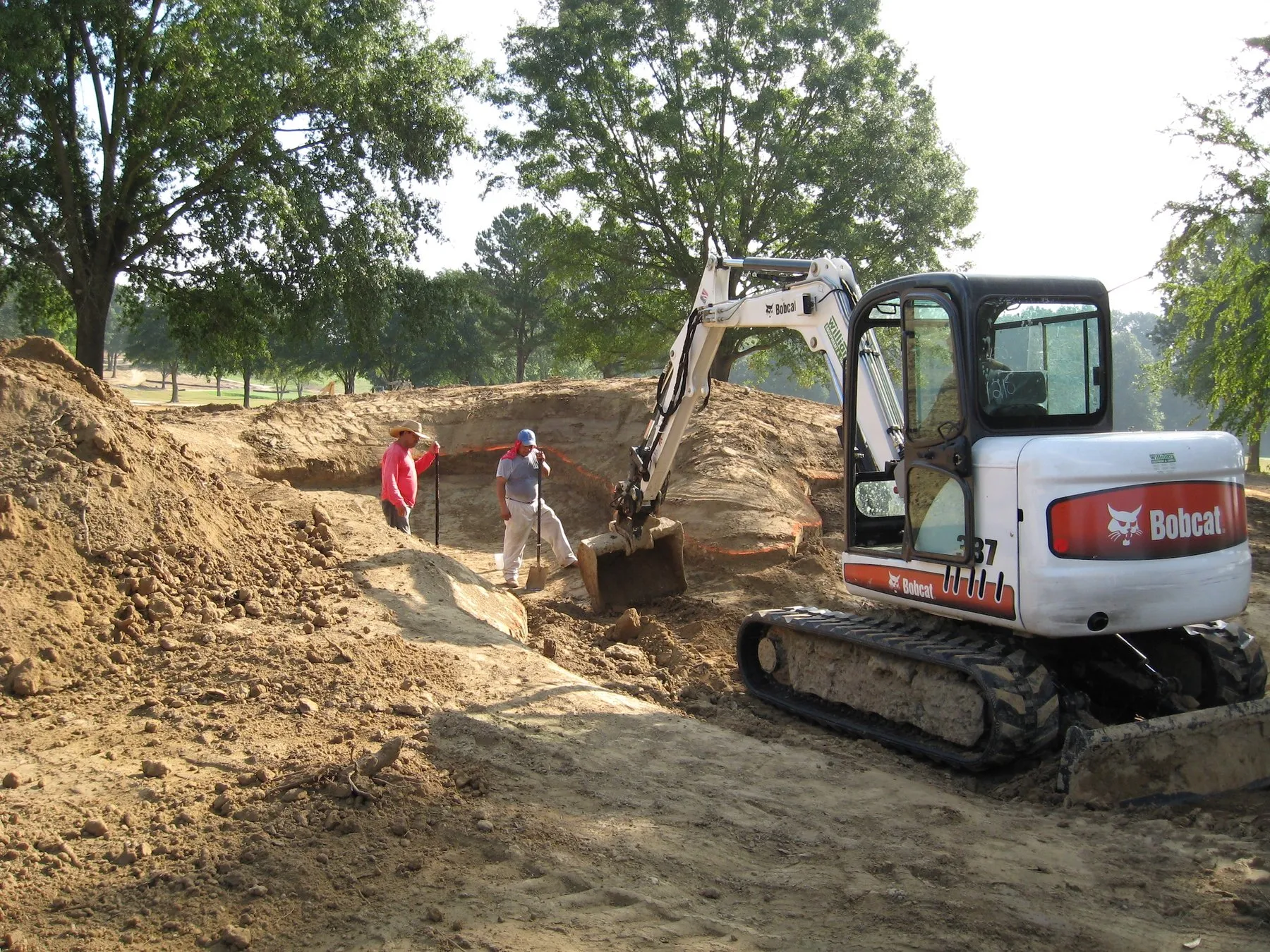 A compact excavator cuts the edge of a new bunker at Mirimichi while two crew members work alongside.
