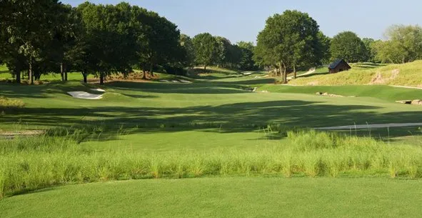 A rolling fairway framed by mature trees, with bunkers and a small building in the distance.