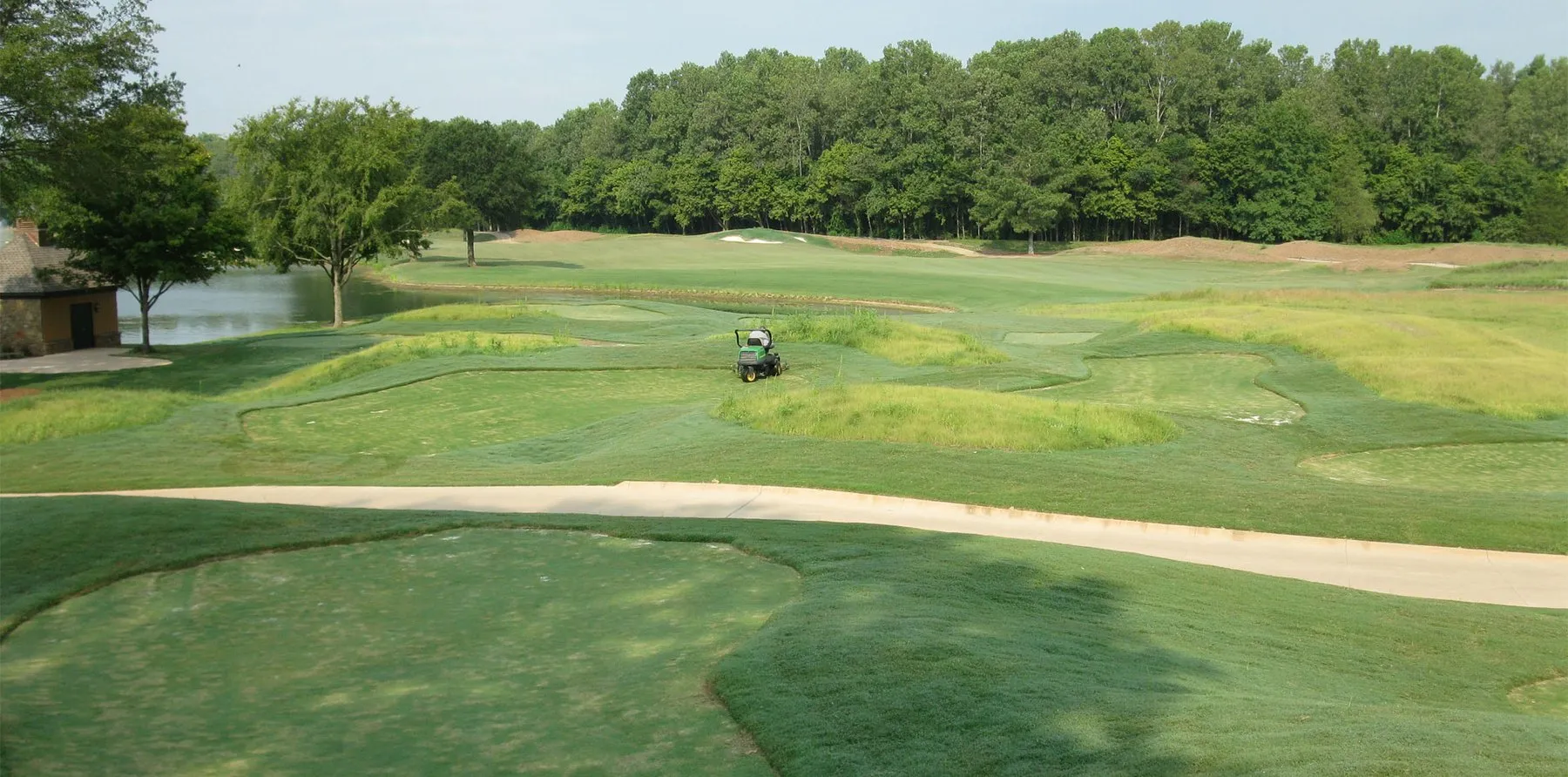 A wide fairway curving past a lake and a tree line, with a golf cart on the cart path.