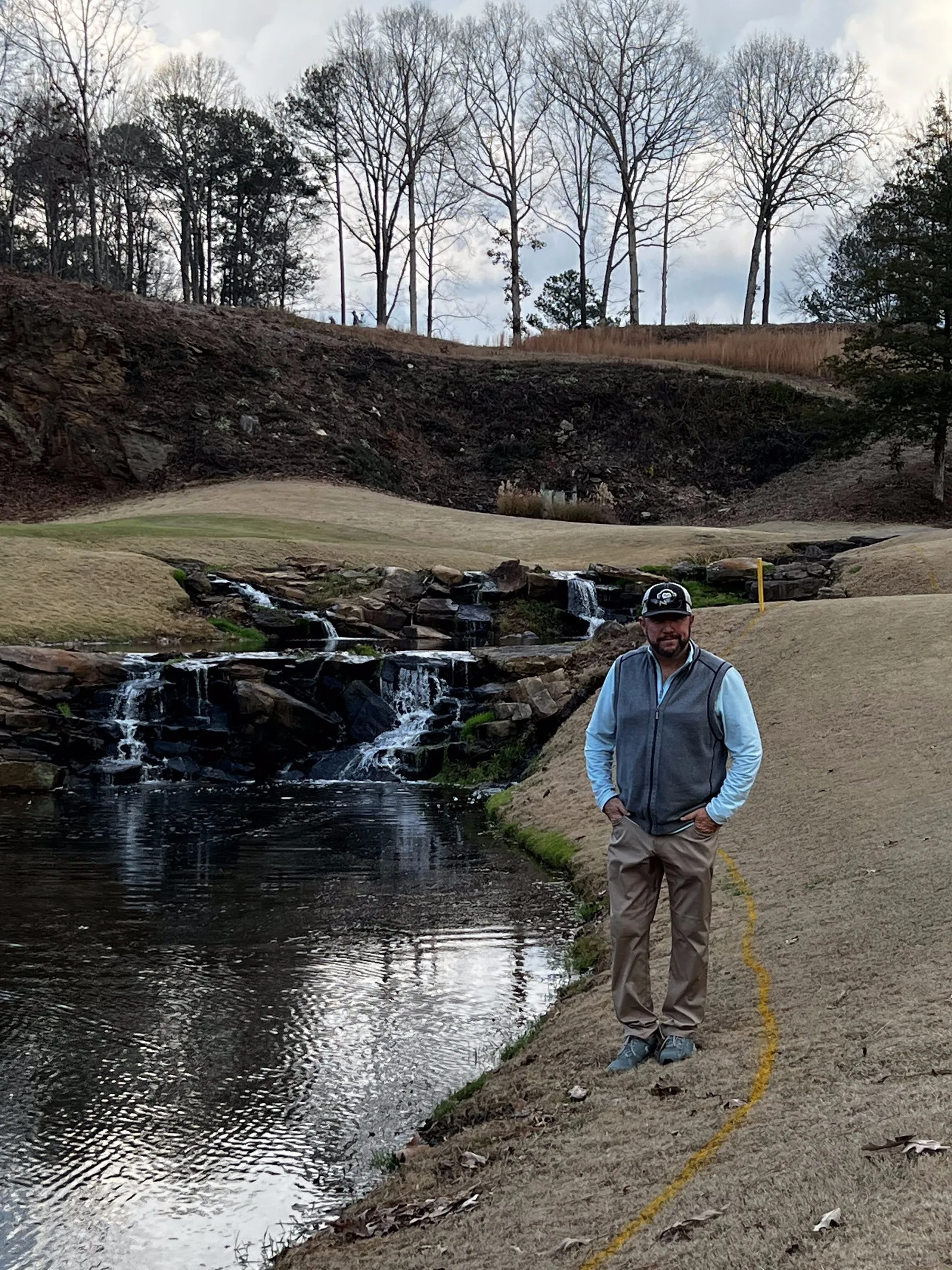 A man stands with his hands in his pockets on the mown bank beside a rock waterfall spilling into a pond, bare winter trees on the ridge behind him.