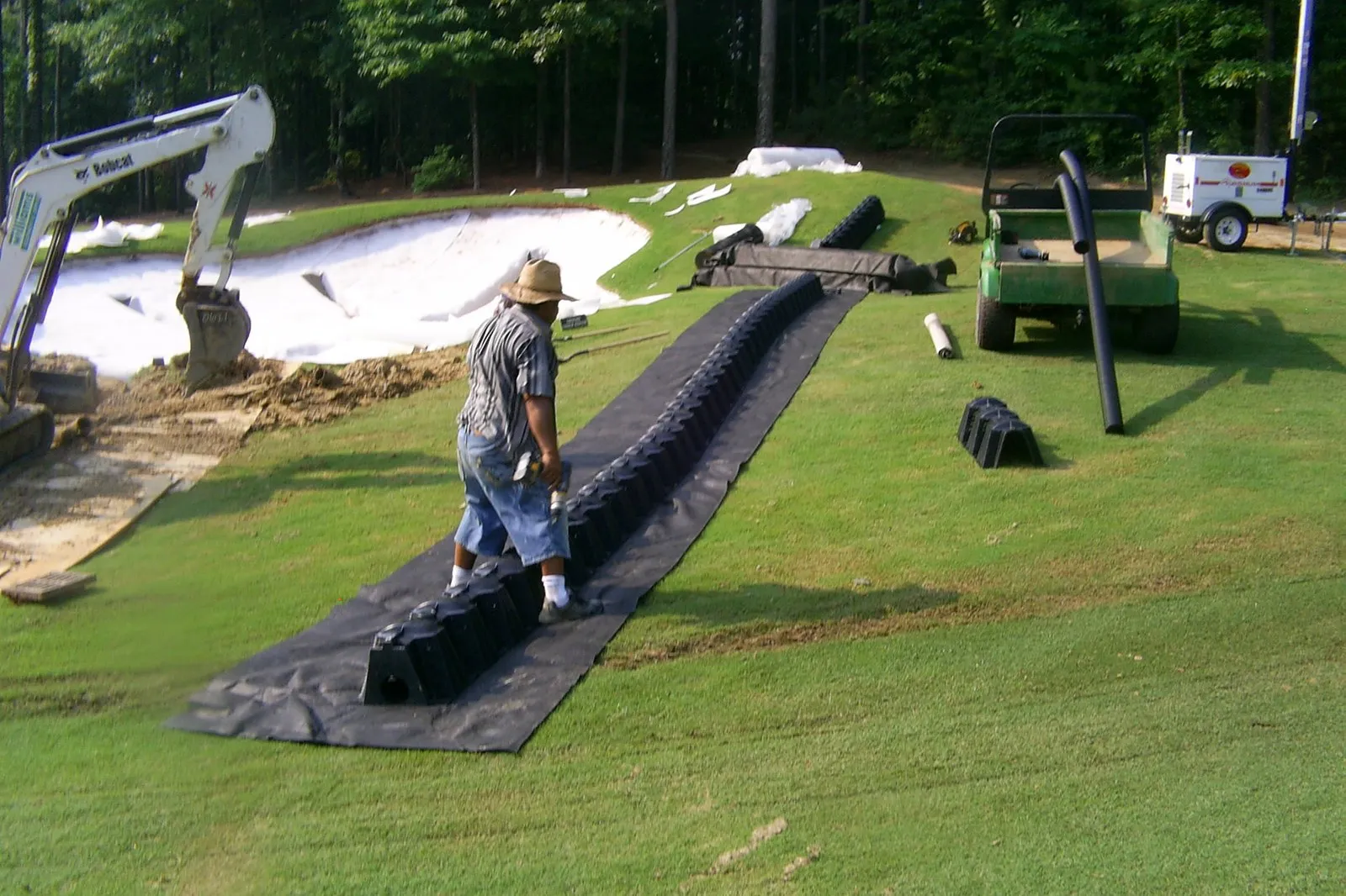 A worker in a straw hat rolls out black drainage pipe across mown turf beside a newly shaped bunker, with a mini excavator behind him.