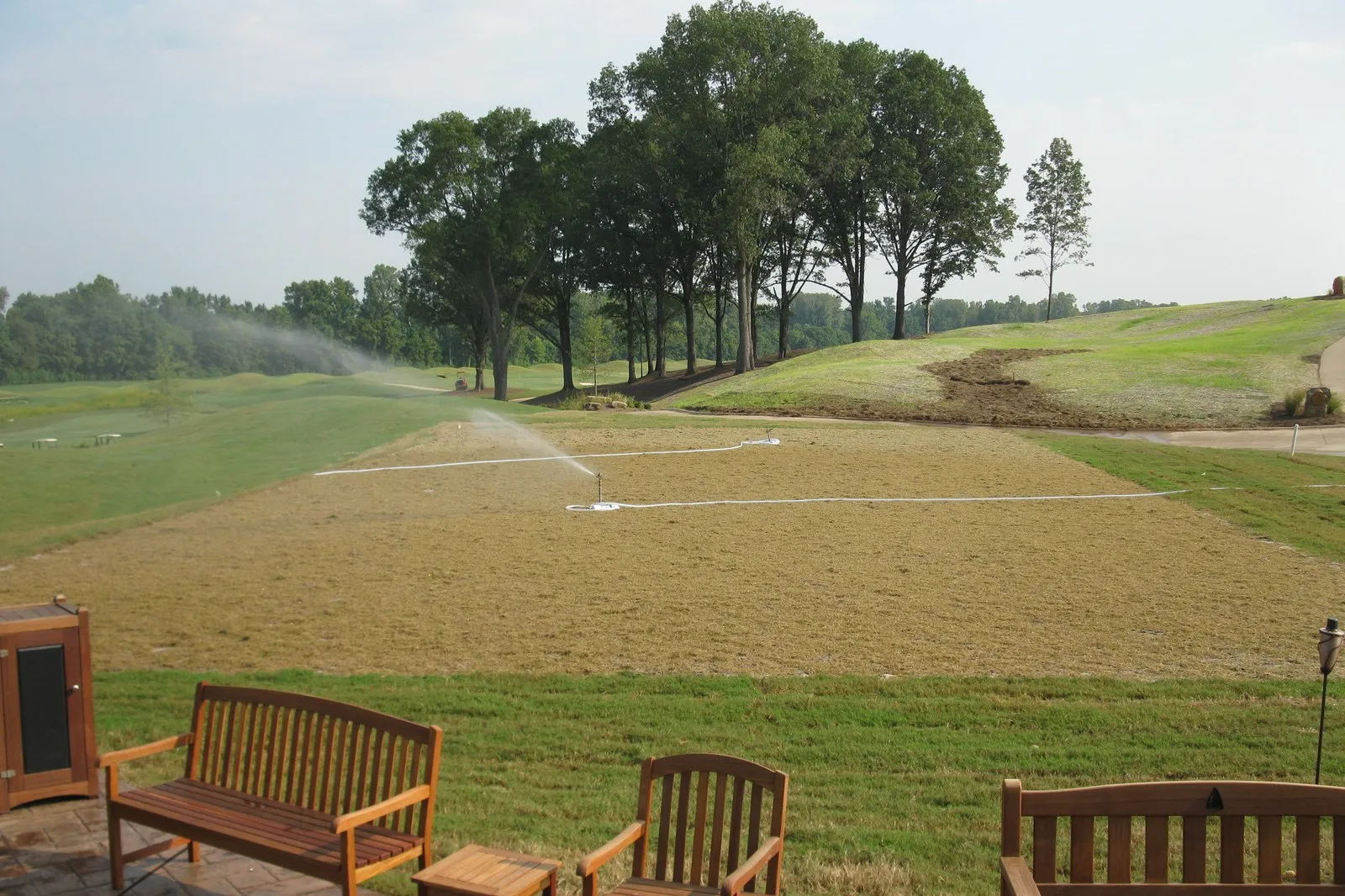 Sprinklers running over newly graded and seeded ground, with a line of mature trees behind.