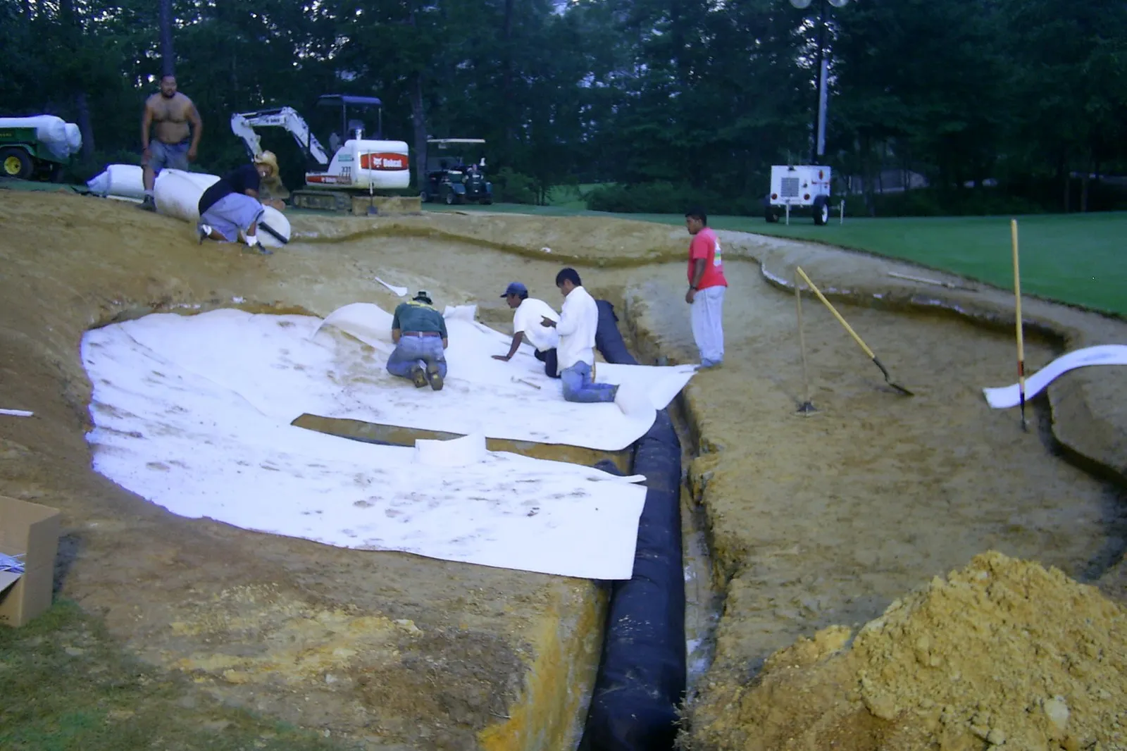 Four crew members kneel in a large bunker under renovation, laying white liner fabric over a drainage trench, with trucks and equipment on the grass behind.