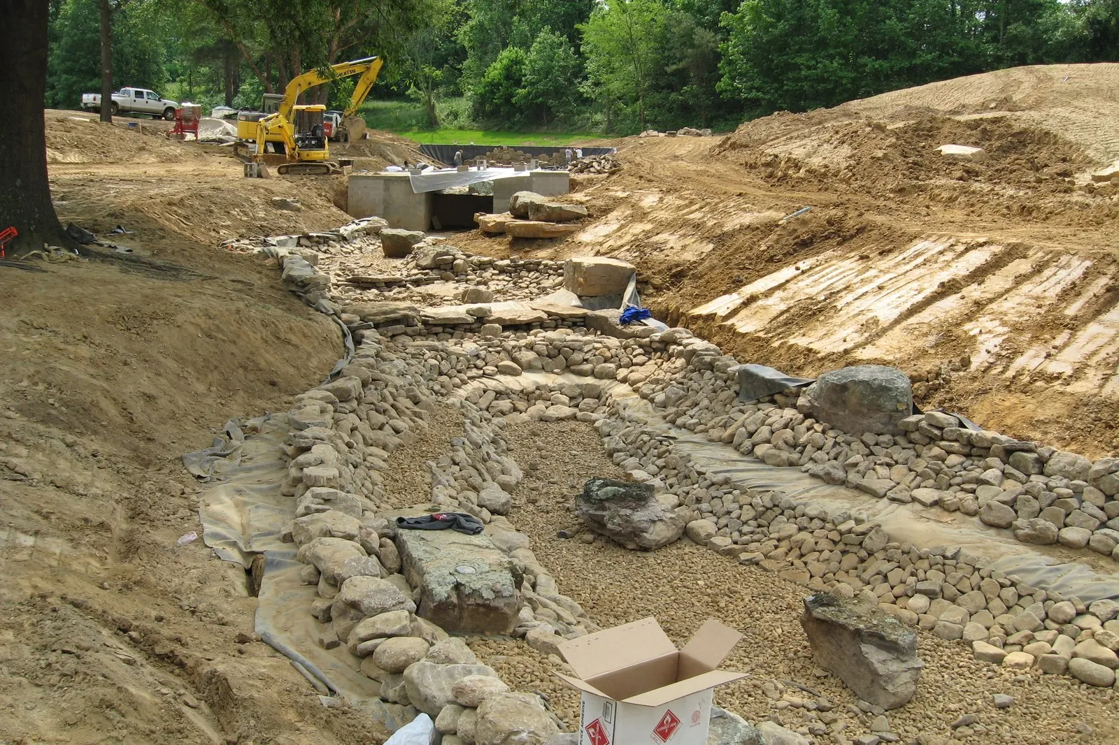 An excavated streambed lined with stacked rock, terraced cut earth on one bank and an excavator working at the top.