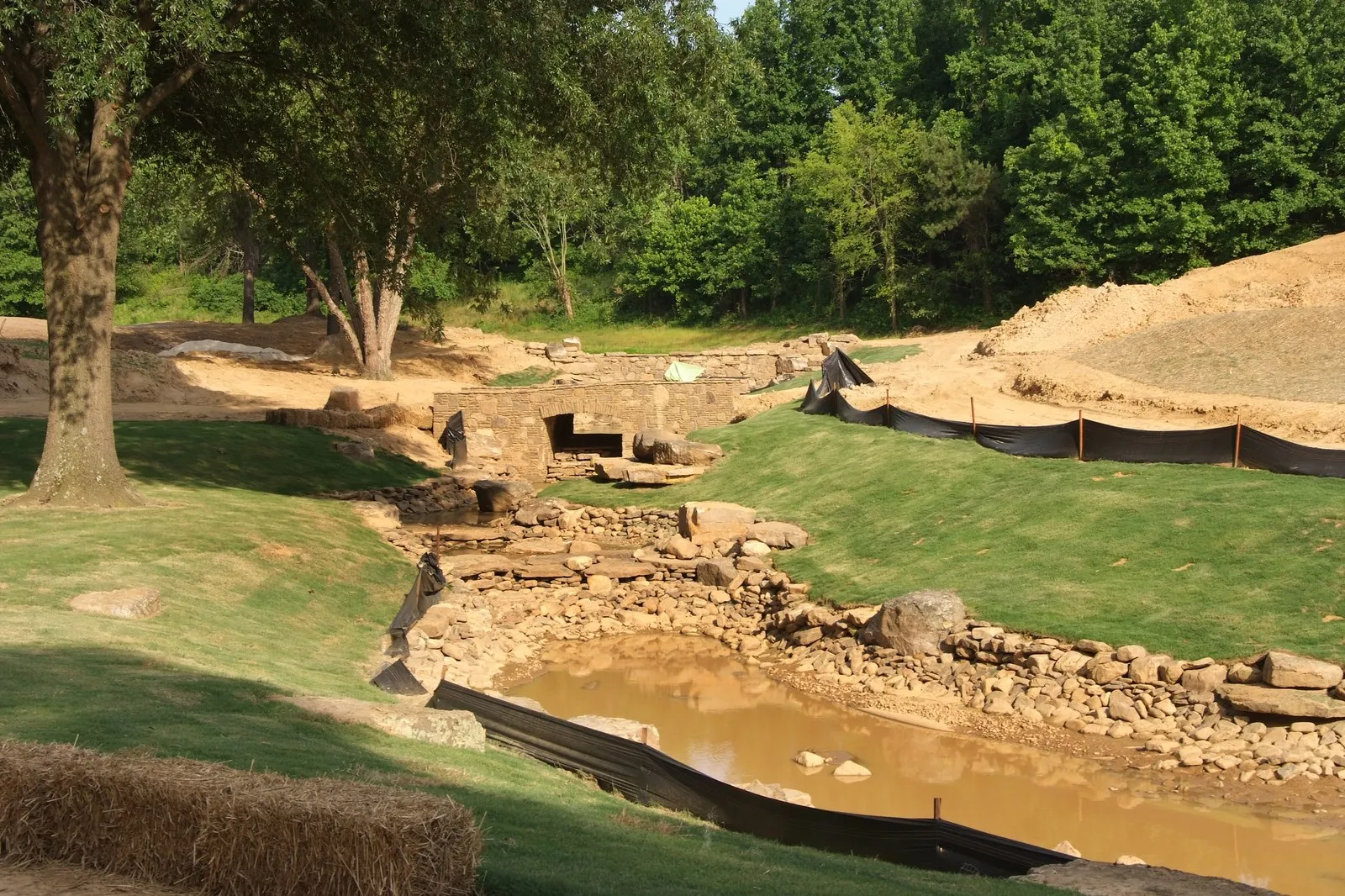 A rock-lined creek under construction running into a muddy pond, straw bales along the bank and mature trees above.
